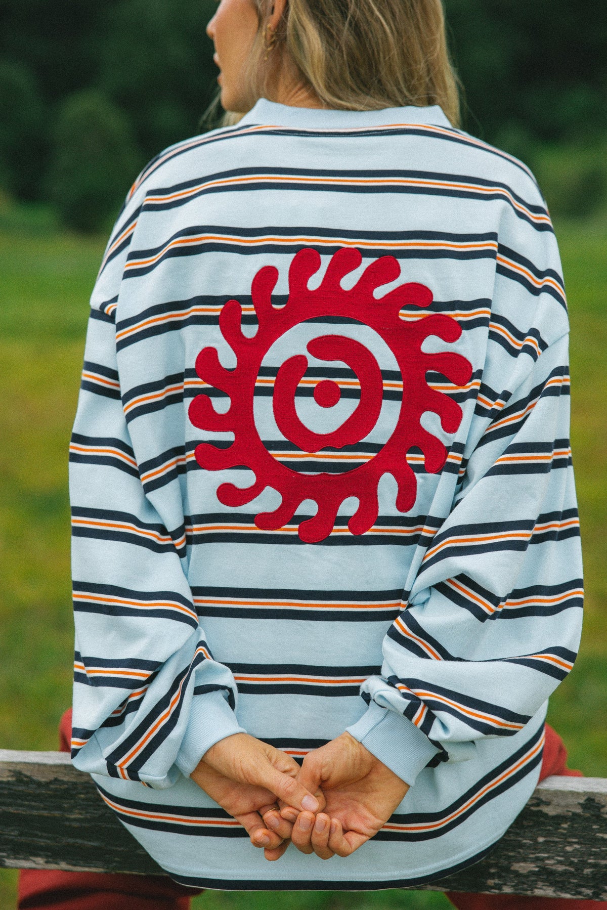 A person viewed from behind wearing a light blue long-sleeve striped shirt with thin navy, orange, and white stripes, featuring a large red circular sun-like graphic on the back. The person is sitting outdoors on a wooden bench with a blurred green natural background.