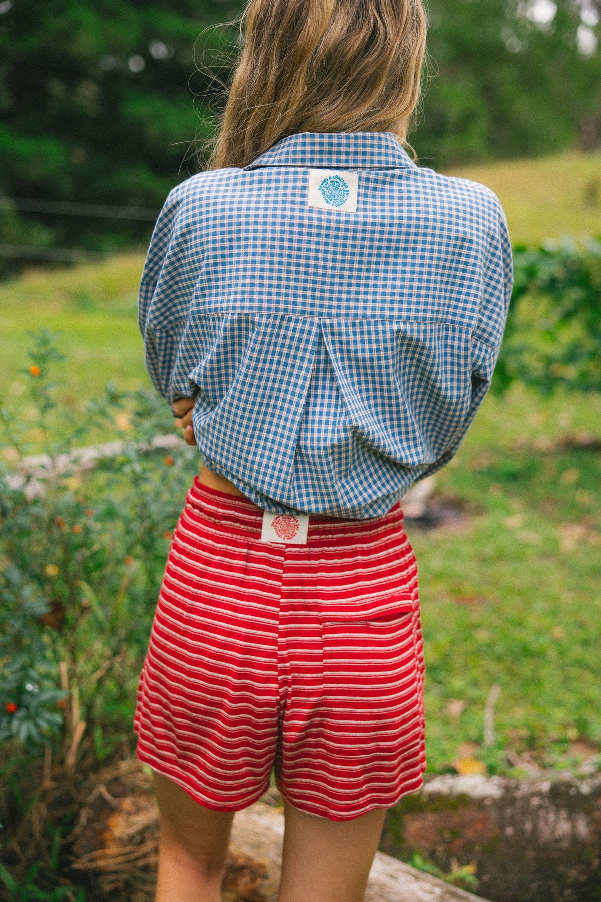 A person standing outdoors in a grassy area, shown from the back, wearing a blue and white gingham button-up shirt and red shorts with thin white horizontal stripes.
