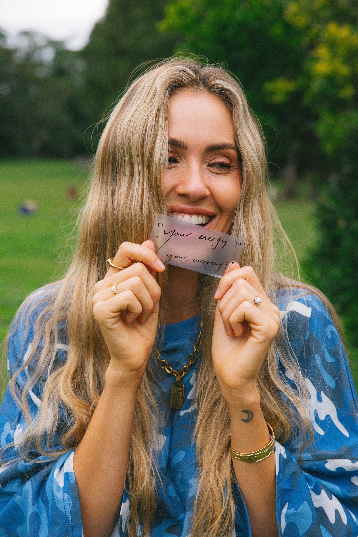 A smiling woman with long blonde hair holds a small card in front of her mouth in an outdoor park setting. She is wearing a blue patterned top and gold jewelry.