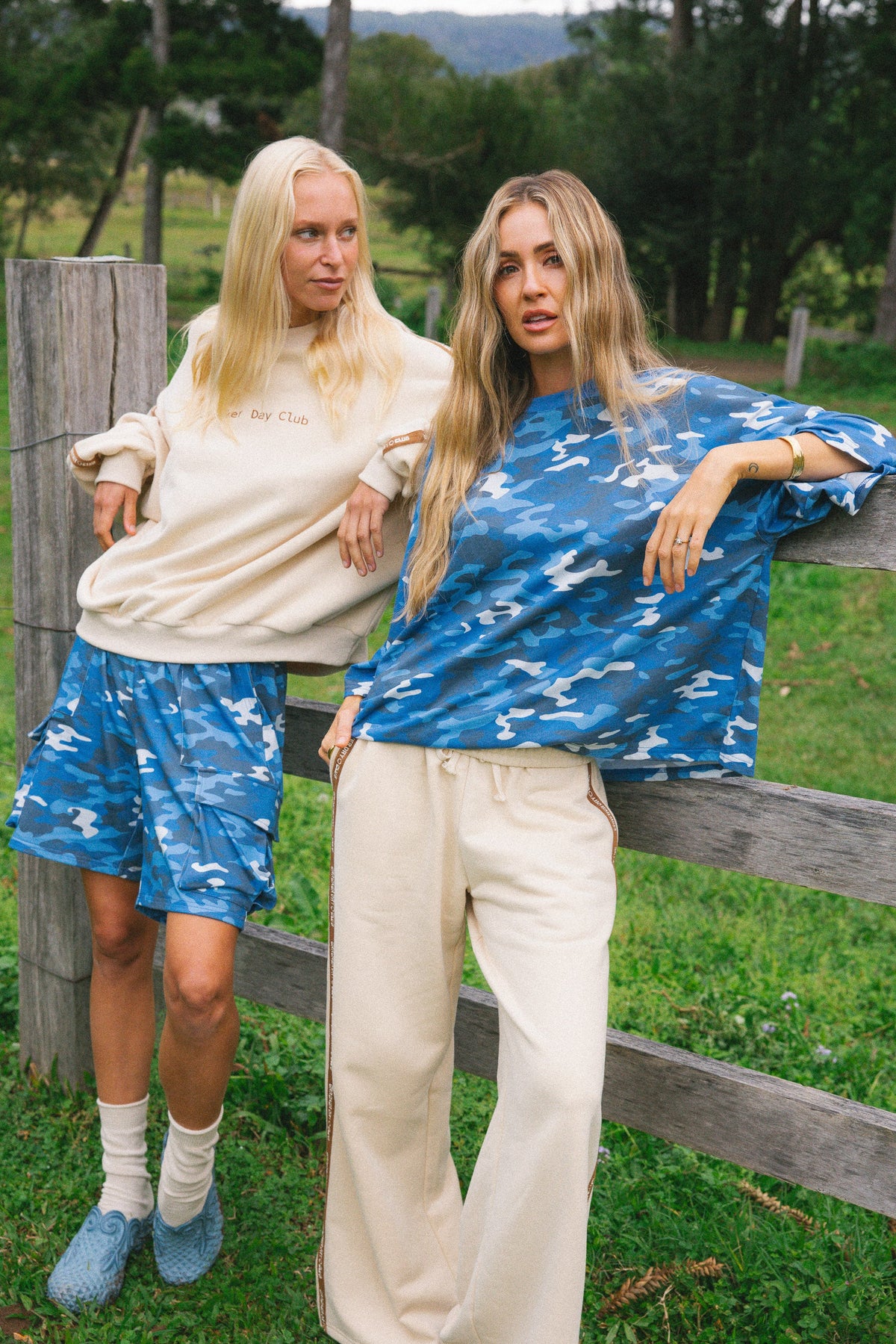 Two women posing outdoors in matching blue and white camouflage lounge sets, with one wearing a cream sweatshirt and shorts and the other wearing a blue patterned top and cream pants, standing behind a wooden fence in a grassy field.