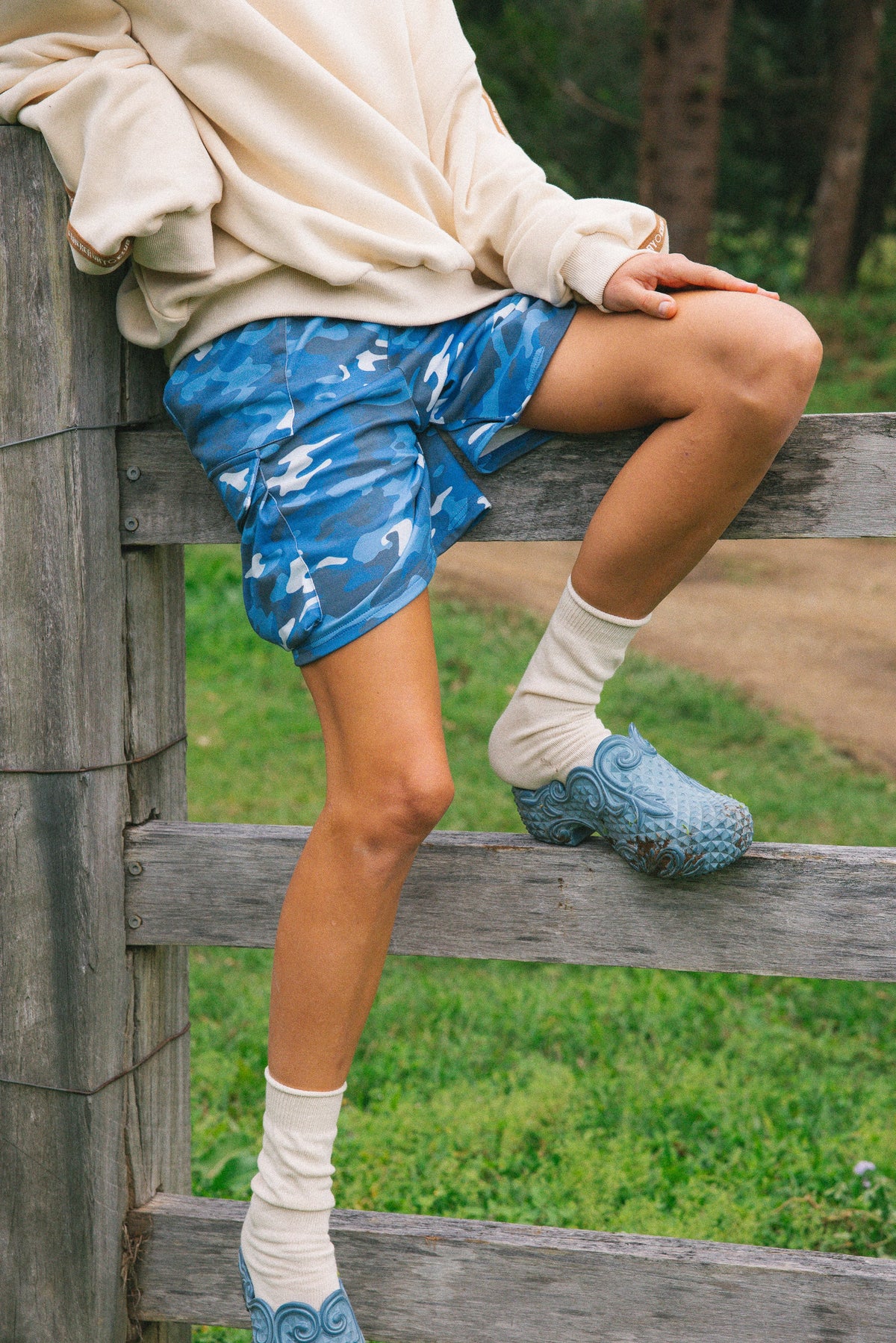 A person outdoors leaning on a wooden fence, wearing blue camouflage-patterned cargo shorts, white crew socks, and light blue clogs.