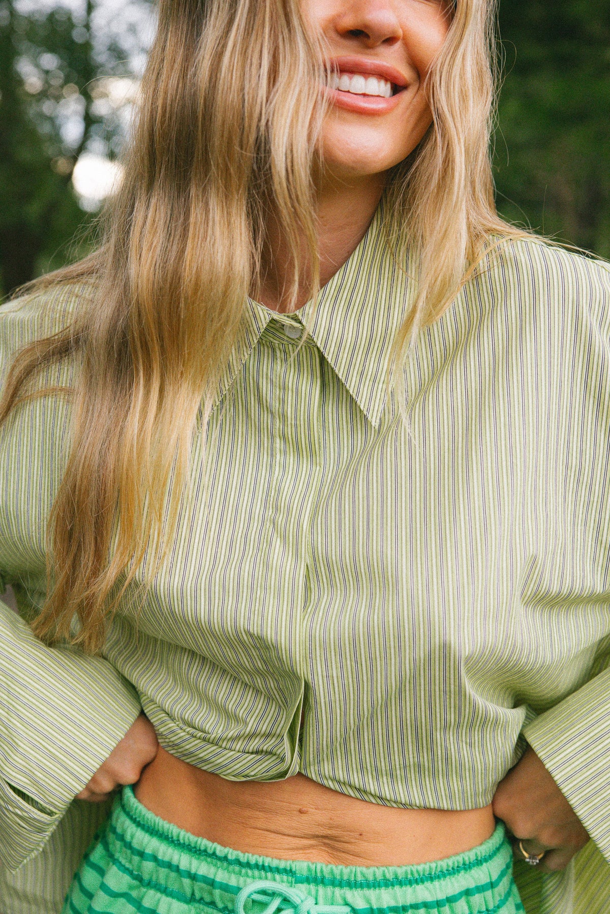 A woman outdoors wearing a light sage green and white vertically striped button-up shirt and bright green drawstring shorts.