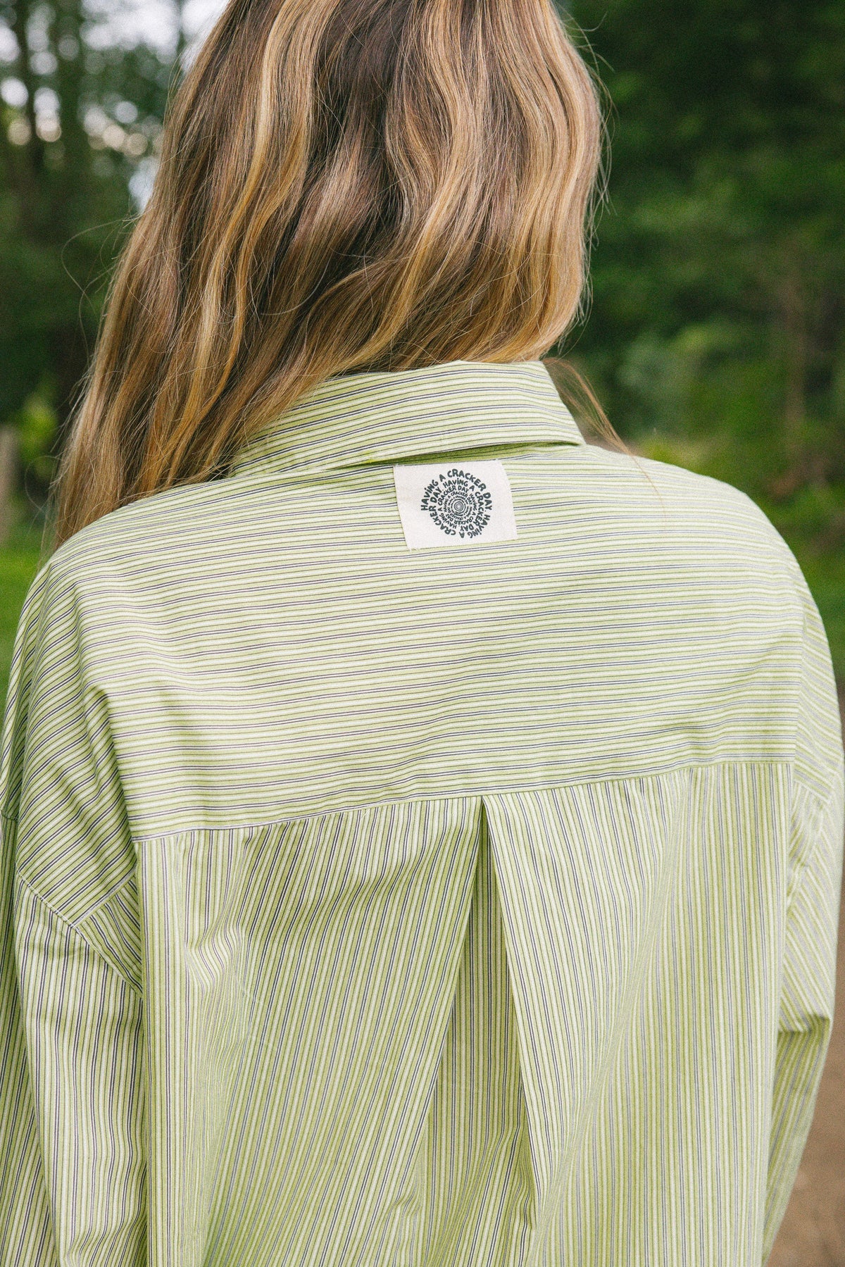 A person viewed from behind wearing a light sage green and white vertically striped button-down shirt, with a circular black-and-white logo patch on the upper back. The background is a soft-focus outdoor green setting.