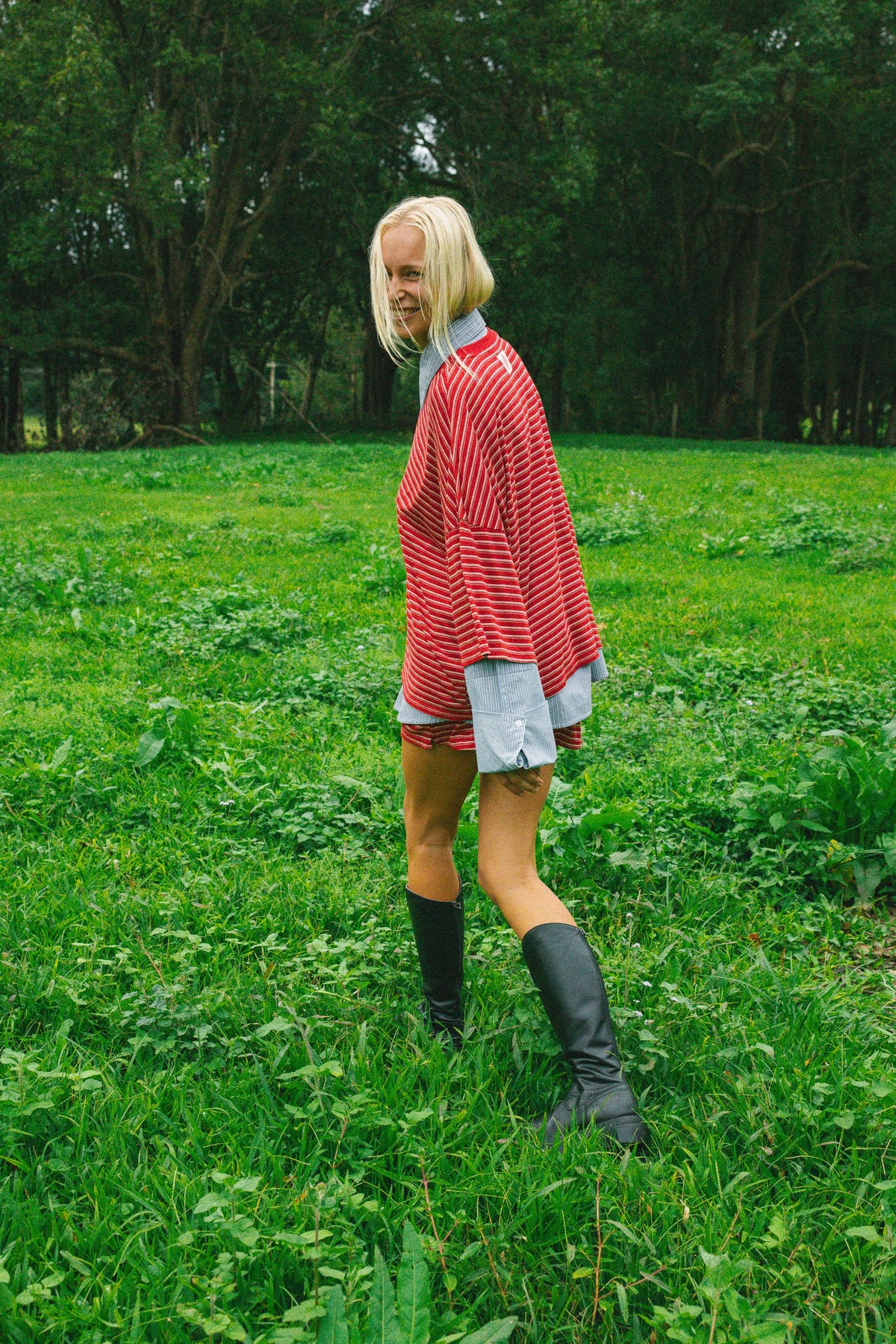A woman standing in a grassy field with trees in the background, wearing a red and white stripedoversize top over a light blue shirt, denim shorts, black knee-high boots, and holding a phone.