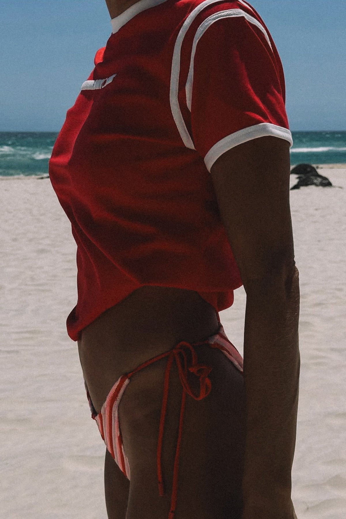 Person wearing a red cracker day club baby tee and bikini bottoms on a beach