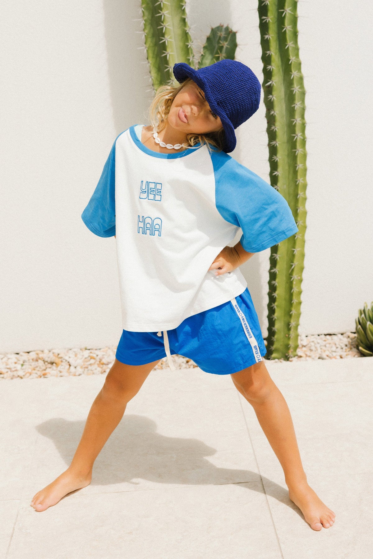 Child wearing a blue and white baseball shirt with yee haa on the front and shorts with a blue cap, standing in front of cacti.