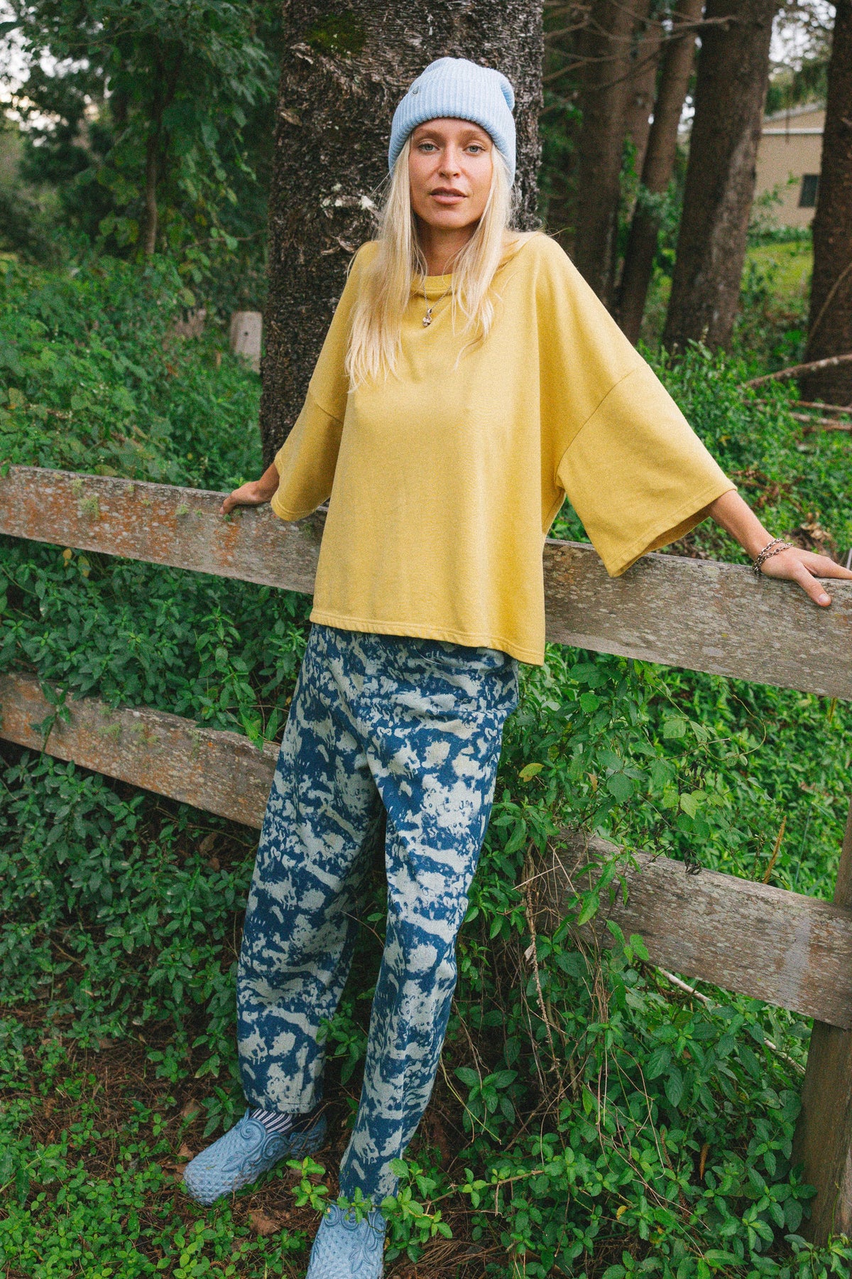 A woman stands outdoors on a dirt path next to a wheelbarrow, wearing a yellow long-sleeve top, blue-and-white patterned jeans, a blue-and-white striped knit scarf, and blue knit shoes.