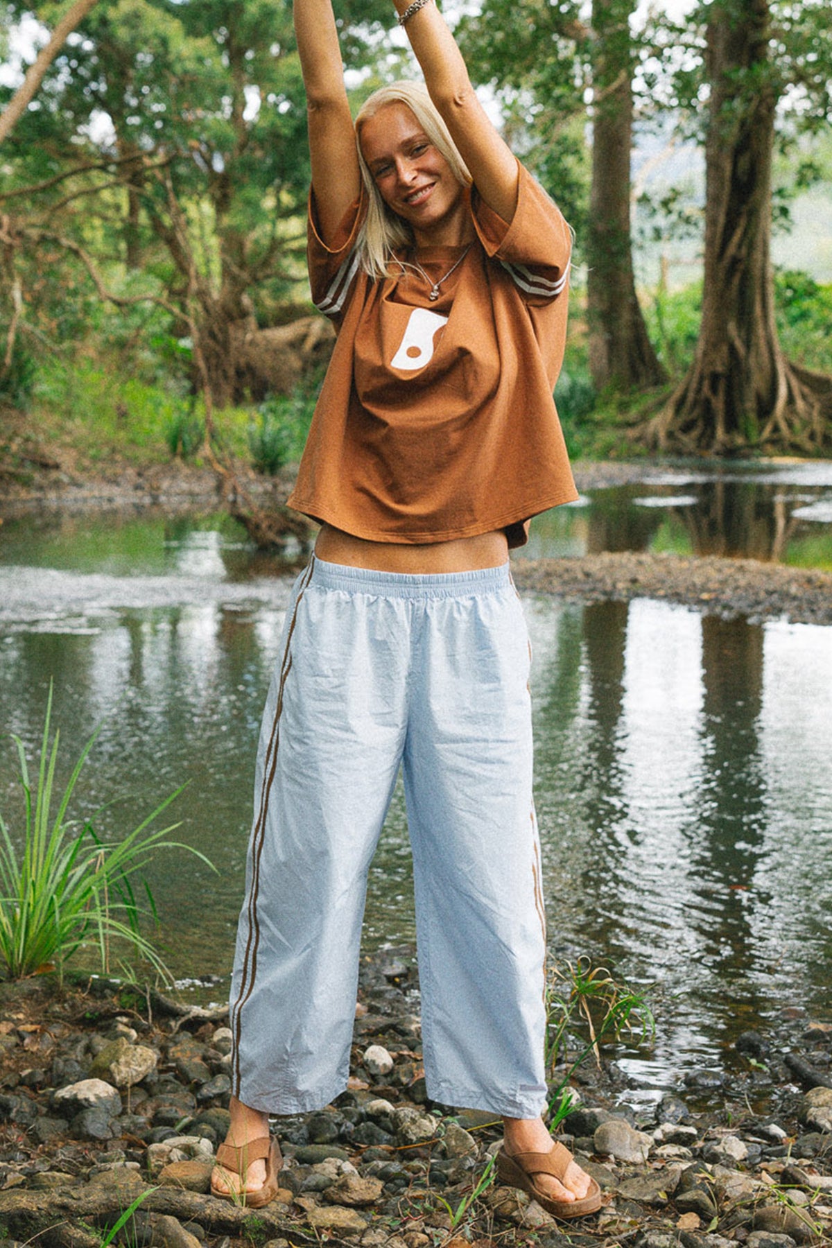 A woman standing outdoors by a river with trees in the background, wearing a brown cropped t-shirt with a white graphic and light blue wide-leg lightweight pants with dark side stripes.