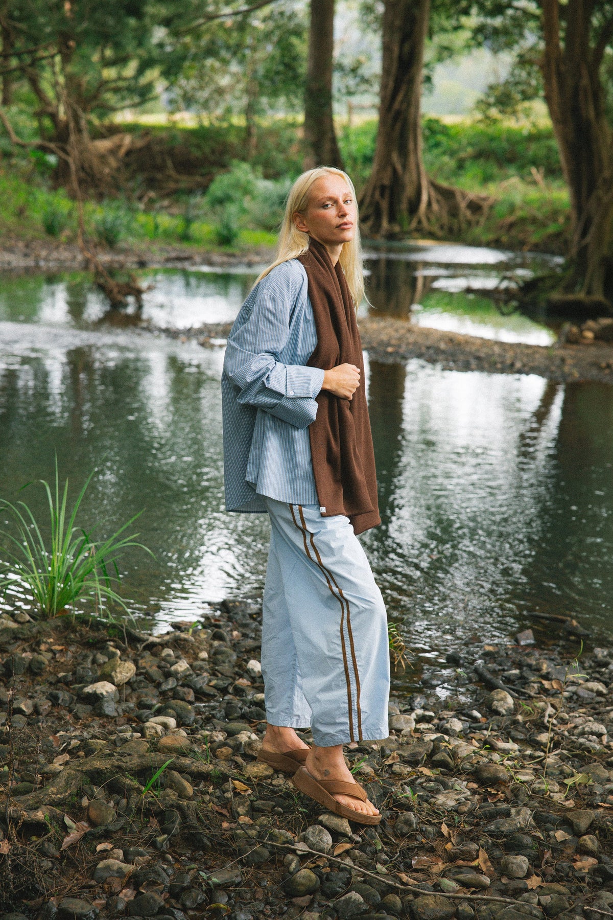 A woman stands on a rocky riverbank in a forest, wearing a light blue button-up shirt, matching light blue wide-leg pants with brown side stripes, a brown wrap, and tan flat shoes.