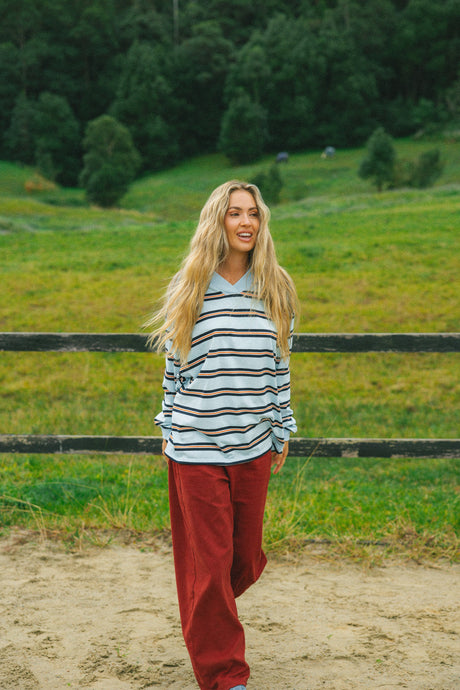 A woman stands outdoors in a grassy field wearing a light blue long-sleeve polo shirt with thin dark horizontal stripes and loose rust-red pants.