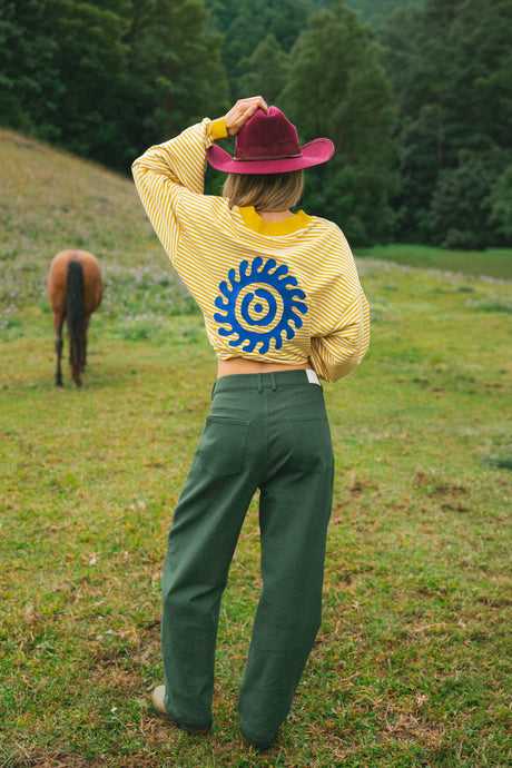 A person standing in a grassy field with trees in the background, wearing a yellow striped cropped sweatshirt with a blue spiral graphic on the back, green pants, and a burgundy hat.