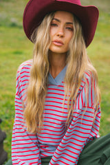 A woman wearing a burgundy wide-brim hat, a red and light blue striped long-sleeve shirt, and dark green pants, sitting outdoors on a grassy field.