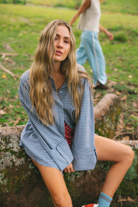 A woman with long blonde hair sits on a mossy log outdoors, wearing an oversized blue and white gingham button-down shirt over a red striped top, paired with light blue socks and red-and-white sneakers. A blurred person walks in the background on a grassy field.