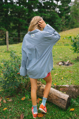 A woman stands outdoors in a grassy yard wearing an oversized light blue and white gingham button-down shirt, red shorts, and orange sneakers.