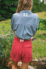 A person standing outdoors in a grassy area, shown from the back, wearing a blue and white gingham button-up shirt and red shorts with thin white horizontal stripes.