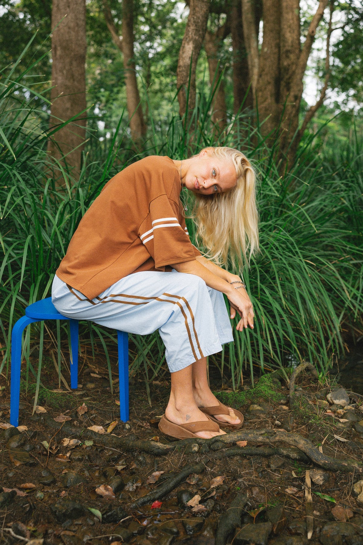 A woman with blonde hair sits slouching on a blue plastic stool outdoors in a wooded area with tall green plants. She is wearing a brown short-sleeve top with white stripes on the sleeves, light blue cropped pants with brown stripes, and brown flat sandals.