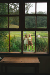 A rustic wooden table in the foreground frames a view through a large multi-pane window of two women standing in a grassy field with trees in the background.
