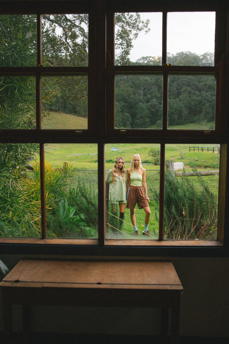 A rustic wooden table in the foreground frames a view through a large multi-pane window of two women standing in a grassy field with trees in the background.
