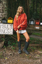 A woman stands outdoors in a wooded area wearing a rust-red sweatshirt with white stripes, white shorts, and black rubber boots. She is posed in front of a rustic wooden structure with small colorful houses and a sign.