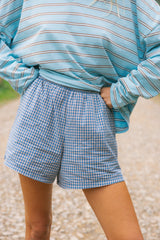 A person wearing blue and white gingham check shorts with a light blue striped long-sleeve top, standing outdoors on a gravel path.