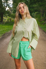 A woman standing on a dirt path in a wooded outdoor setting, wearing a light green vertically striped button-up shirt and green-and-white horizontally striped shorts.