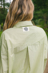 A person viewed from behind wearing a light sage green and white vertically striped button-down shirt, with a circular black-and-white logo patch on the upper back. The background is a soft-focus outdoor green setting.