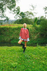A woman stands in a grassy outdoor setting near a small stream, wearing a red-and-white striped poncho-style top with light blue cuffs, a matching striped skirt, and black rain boots.