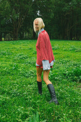 A woman standing in a grassy field with trees in the background, wearing a red and white stripedoversize top over a light blue shirt, denim shorts, black knee-high boots, and holding a phone.