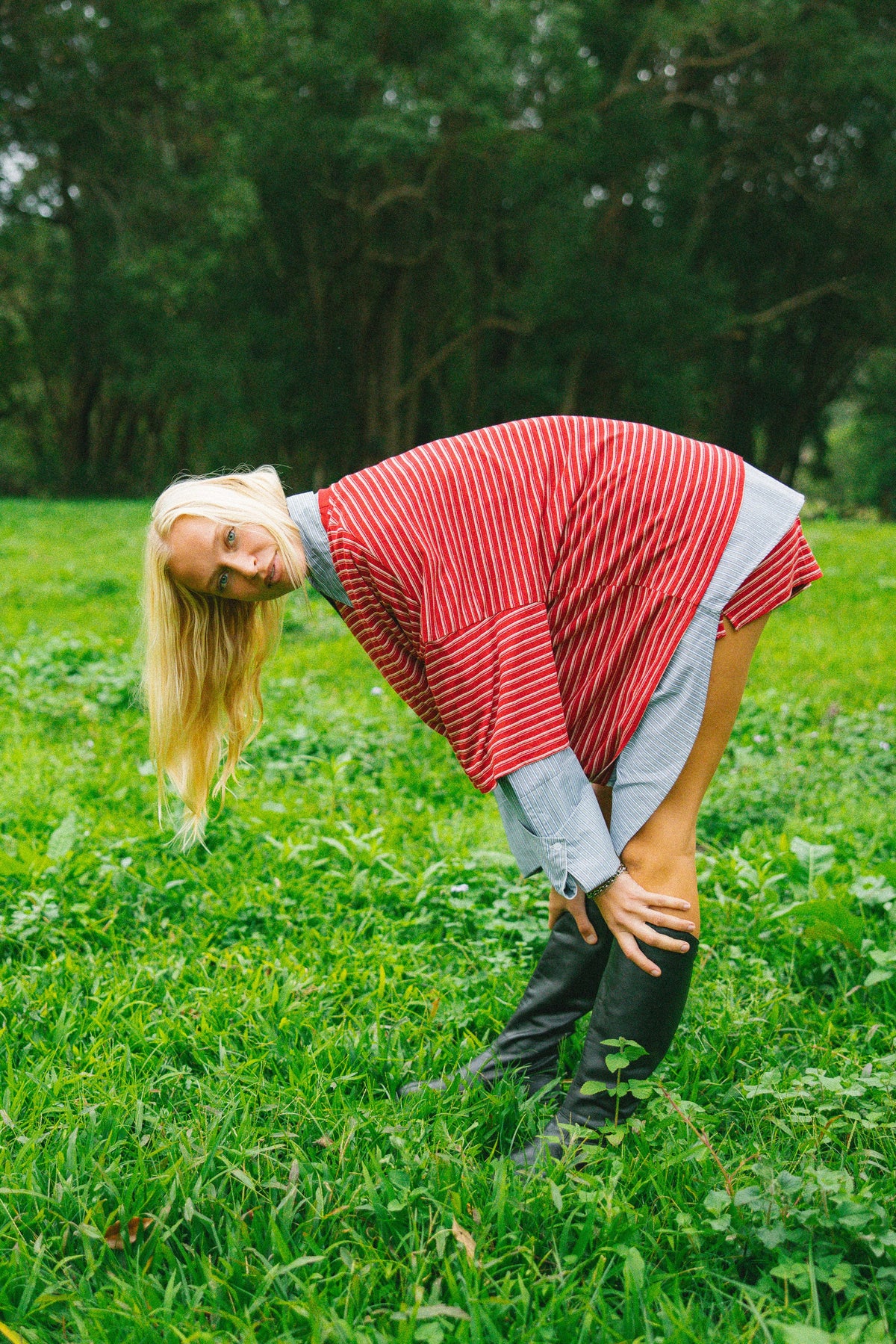 A woman in a red and white striped oversized tee, gray shirt, and black boots bends over in a grassy field with trees in the background.