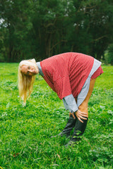 A woman in a red and white striped oversized tee, gray shirt, and black boots bends over in a grassy field with trees in the background.