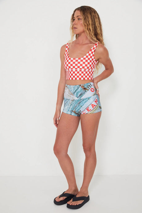 Woman wearing a red and white checkered punchy surf top and patterned blue and white original betty bottoms swim shorts on a white background