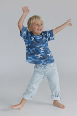 A young blonde girl wearing a blue camouflage t-shirt and light wash jeans, posing with his arms outstretched on a seamless light gray studio background.
