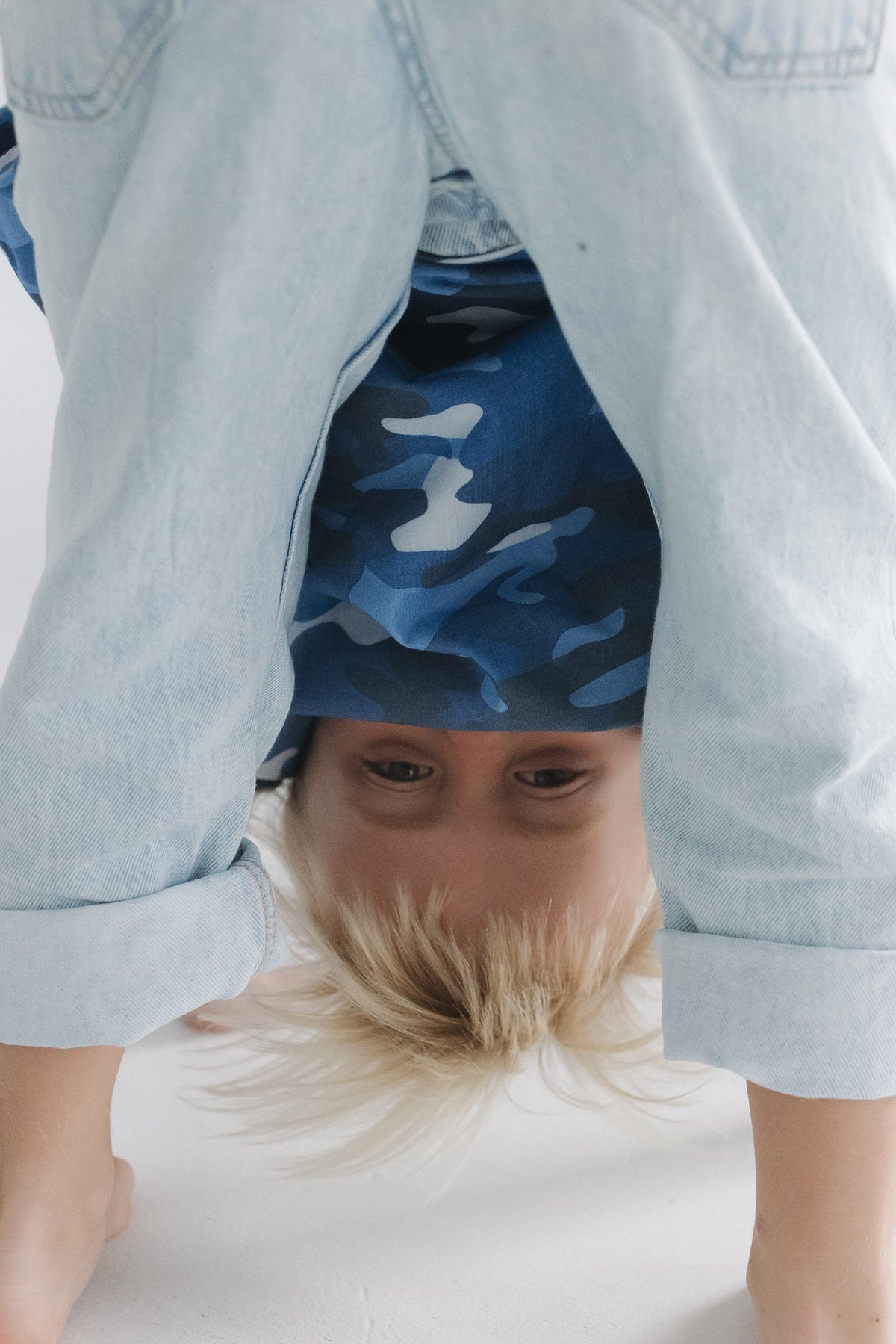 A child wearing a blue and white camouflage t-shirt under a light-wash denim shirt, performing a handstand on a white floor.