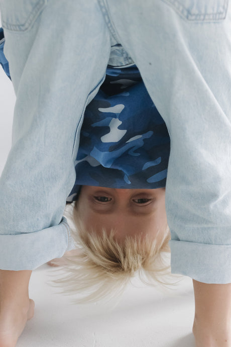 A child wearing a blue and white camouflage t-shirt under a light-wash denim shirt, performing a handstand on a white floor.