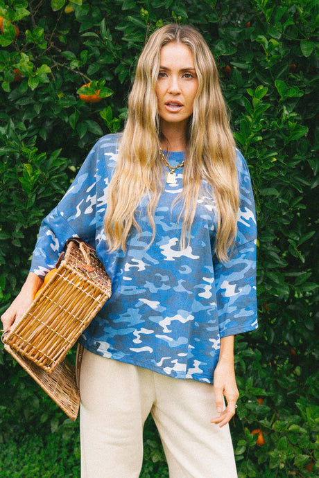 A woman with long blonde hair stands outdoors in front of a green hedge, wearing a blue and white camouflage patterned top, cream pants, and holding a woven straw bag.