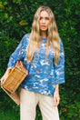 A woman with long blonde hair stands outdoors in front of a green hedge, wearing a blue and white camouflage patterned top, cream pants, and holding a woven straw bag.