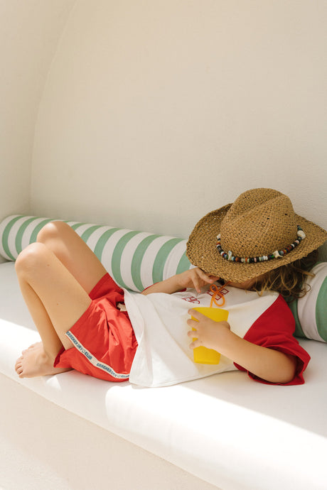 Child wearing a straw hat and red shorts lying on a white couch with green striped cushions.