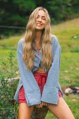 A woman with long blonde hair sits outdoors on a tree trunk, wearing a light blue and white gingham button-up shirt with wide flared sleeves and red-and-white horizontally striped shorts.