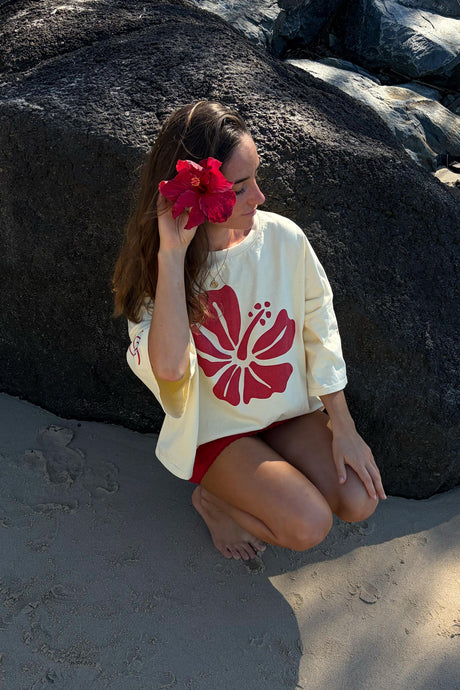 A woman crouches on a sandy beach in front of large dark boulders, wearing a cream short-sleeve top with a large red hibiscus flower graphic and red shorts, while holding a red flower to her eye.