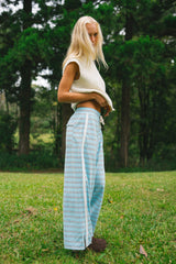 A blonde woman stands on a grassy lawn in front of a forest, wearing a cream sleeveless knit top and light blue wide-leg striped pants.