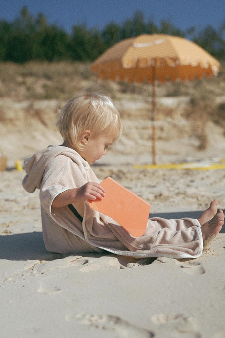 toddler wearing a retro hoodie towel by cracker day club while sitting in the sand on the beach playing with beach toys