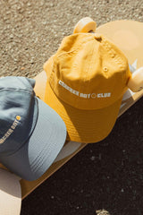 two cracker day club wordmark dad caps in must and blue sitting on a skateboard in the sun