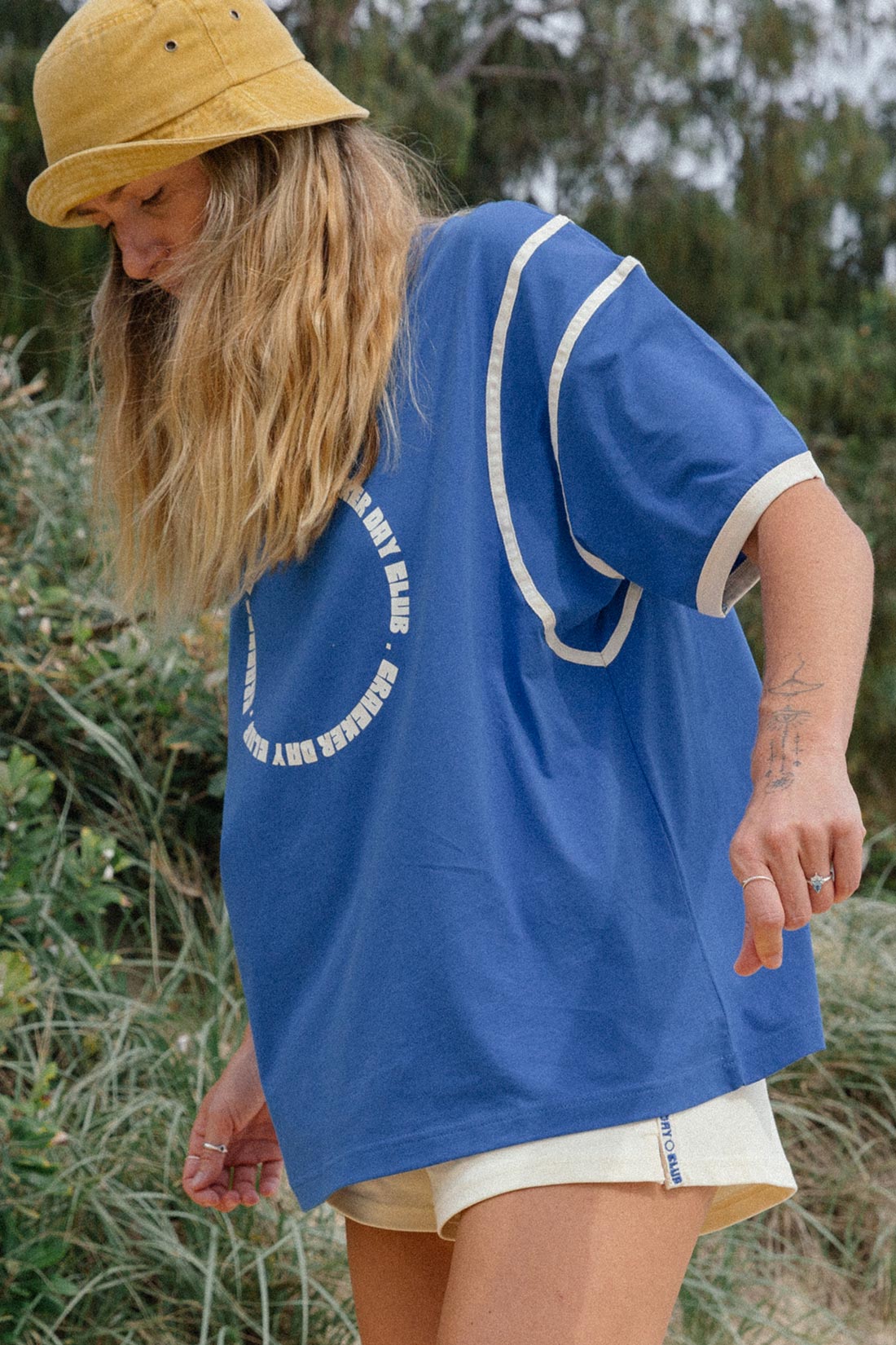 woman at beach wearing the Racer Club Tee by cracker day club in blue with a yellow hat