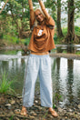 A woman standing outdoors by a river with trees in the background, wearing a brown cropped t-shirt with a white graphic and light blue wide-leg lightweight pants with dark side stripes.