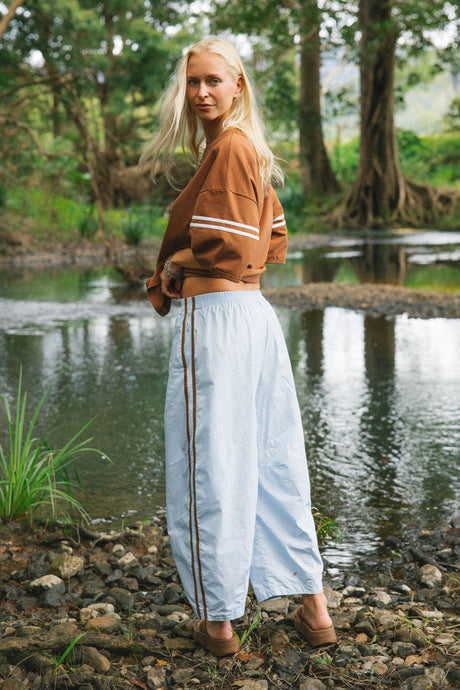 A woman stands outdoors by a river in a wooded area, wearing a brown cropped shirt with white stripes on the sleeves and light blue wide-leg pants with dark side stripes.