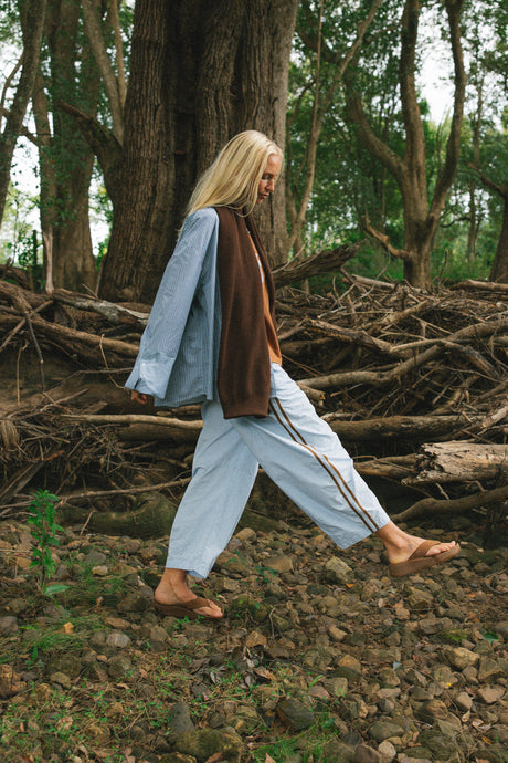 A woman walking through a wooded forest wearing a light blue oversized shirt, a brown vest, light blue wide-leg pants with thin dark stripes, and brown sandals.