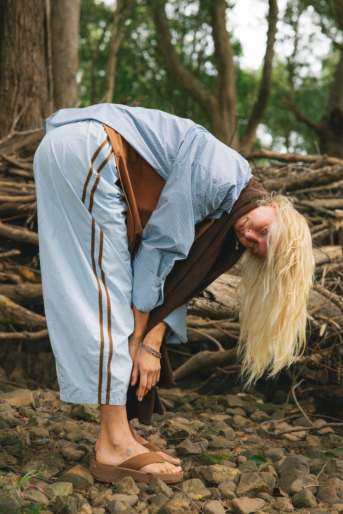 A woman in a light blue hooded sweatshirt and matching light blue pants with brown side stripes, bending forward on a rocky forest path.