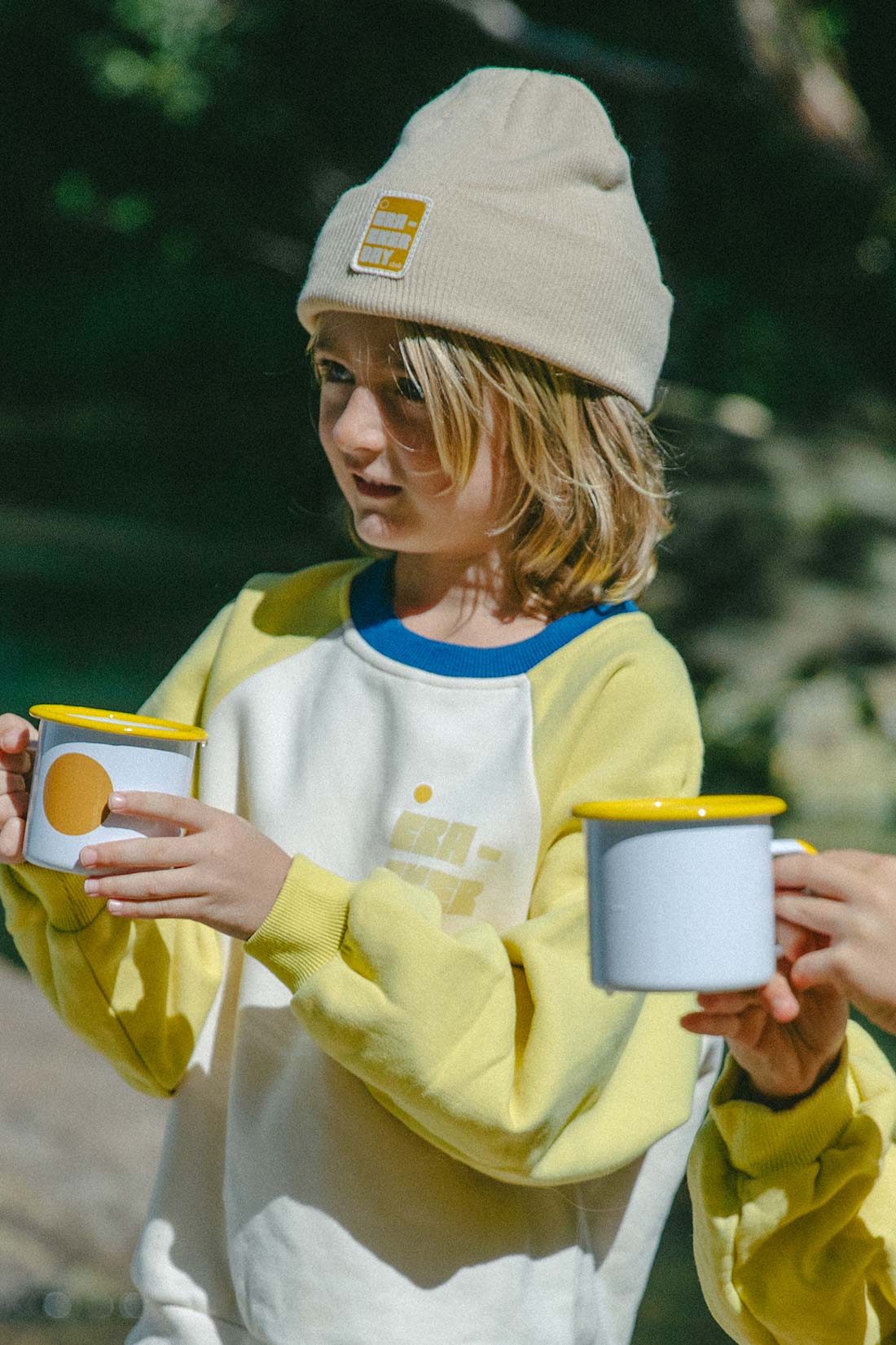 boy with long hair wearing a cracker day beanie while camping out in the rainforest. he is drinking from a camping cup with yellow rim and circle design