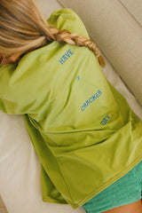 Person wearing a green jacket with have a cracker day text on the back, sitting on a beige couch.